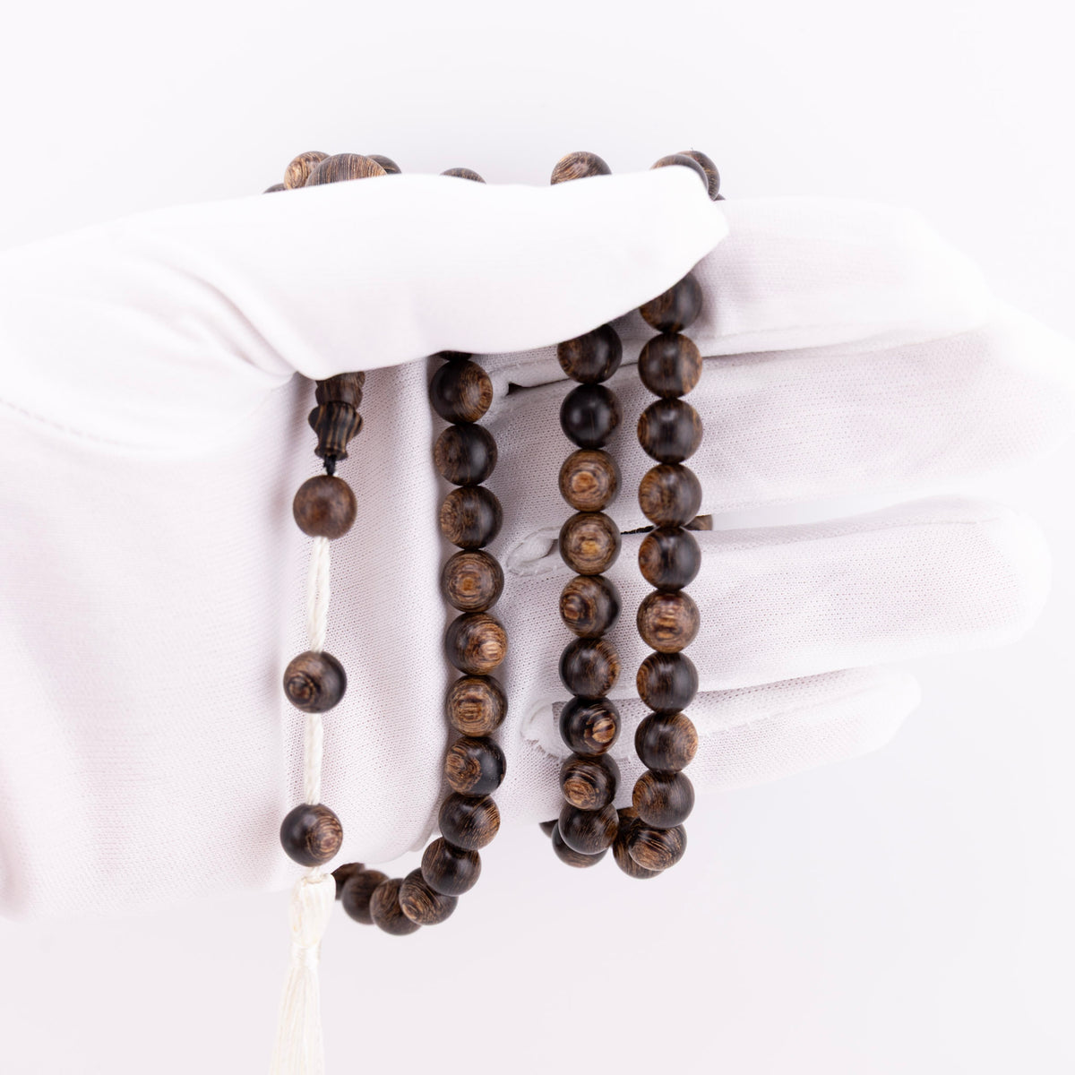 Long agarwood prayer beads with white cord and tassel held in a white-gloved hand